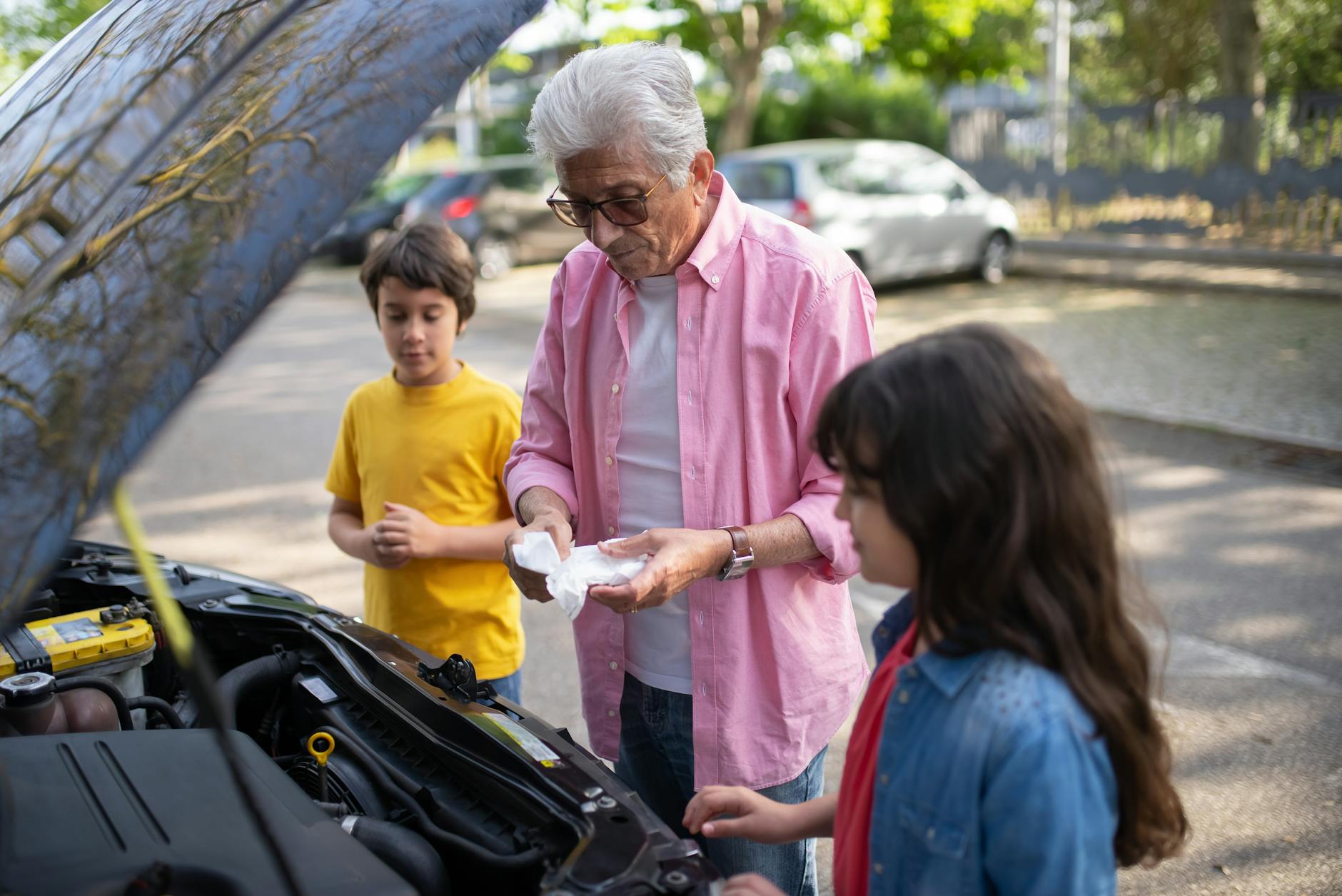 Elderly man shows grandchildren how to care for a car engine outdoors.