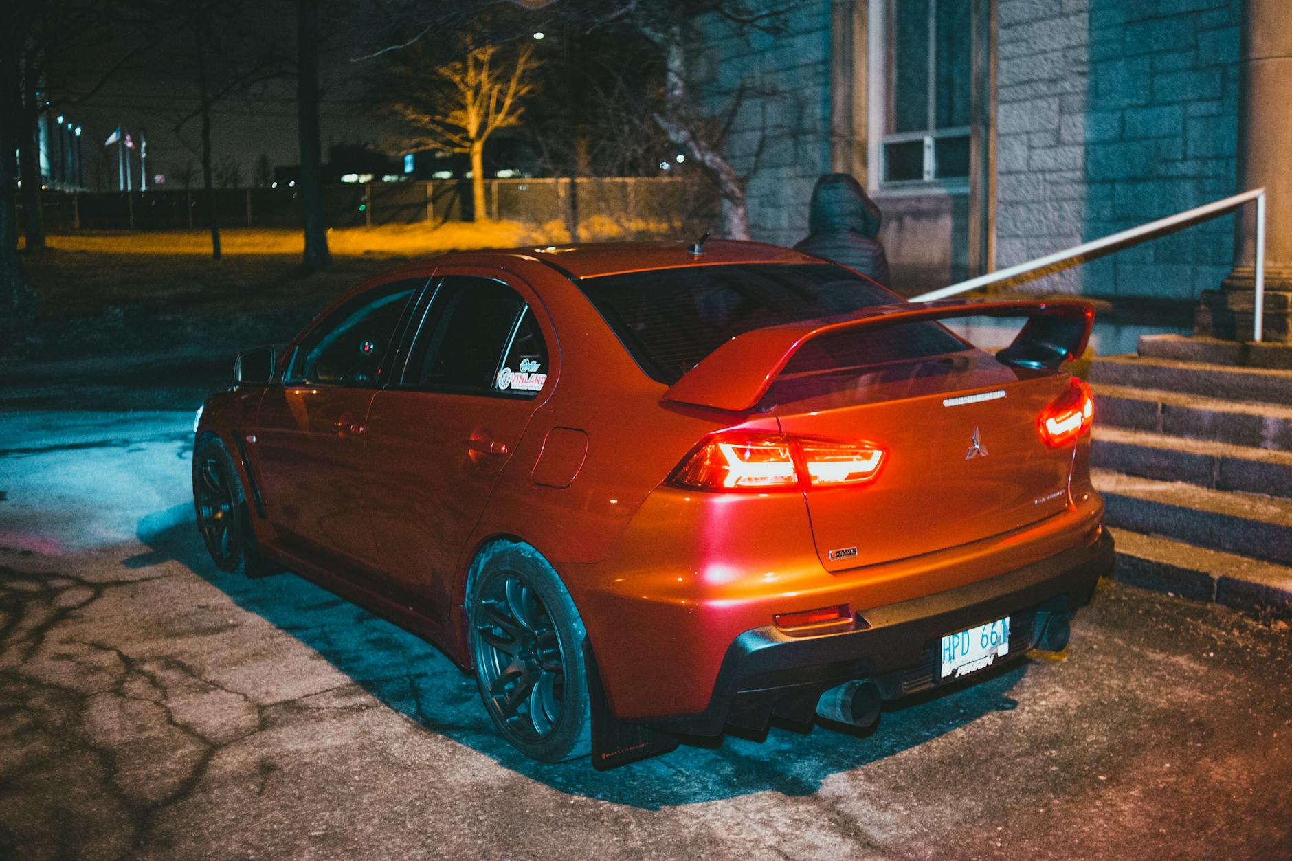 Red modern car parked on asphalt road in town street near building with faceless person near steps at night near trees