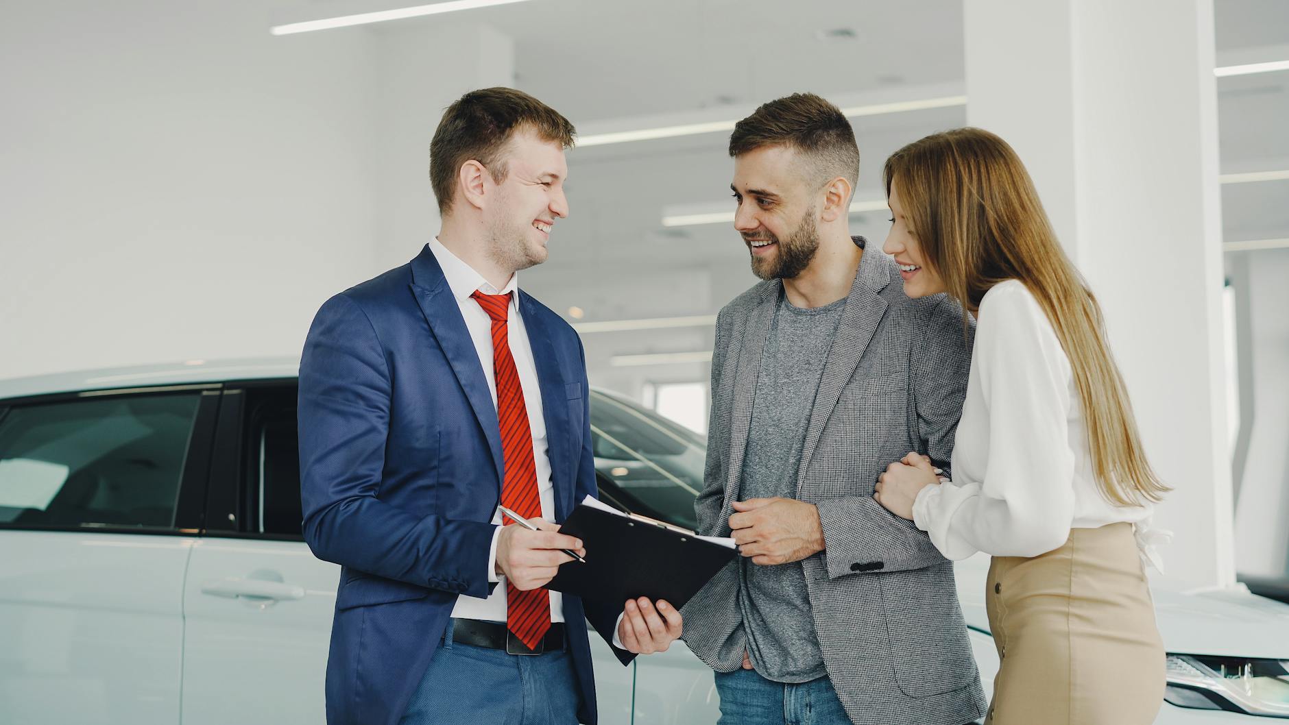 A smiling couple buys a new car from a confident salesman inside a modern car dealership.