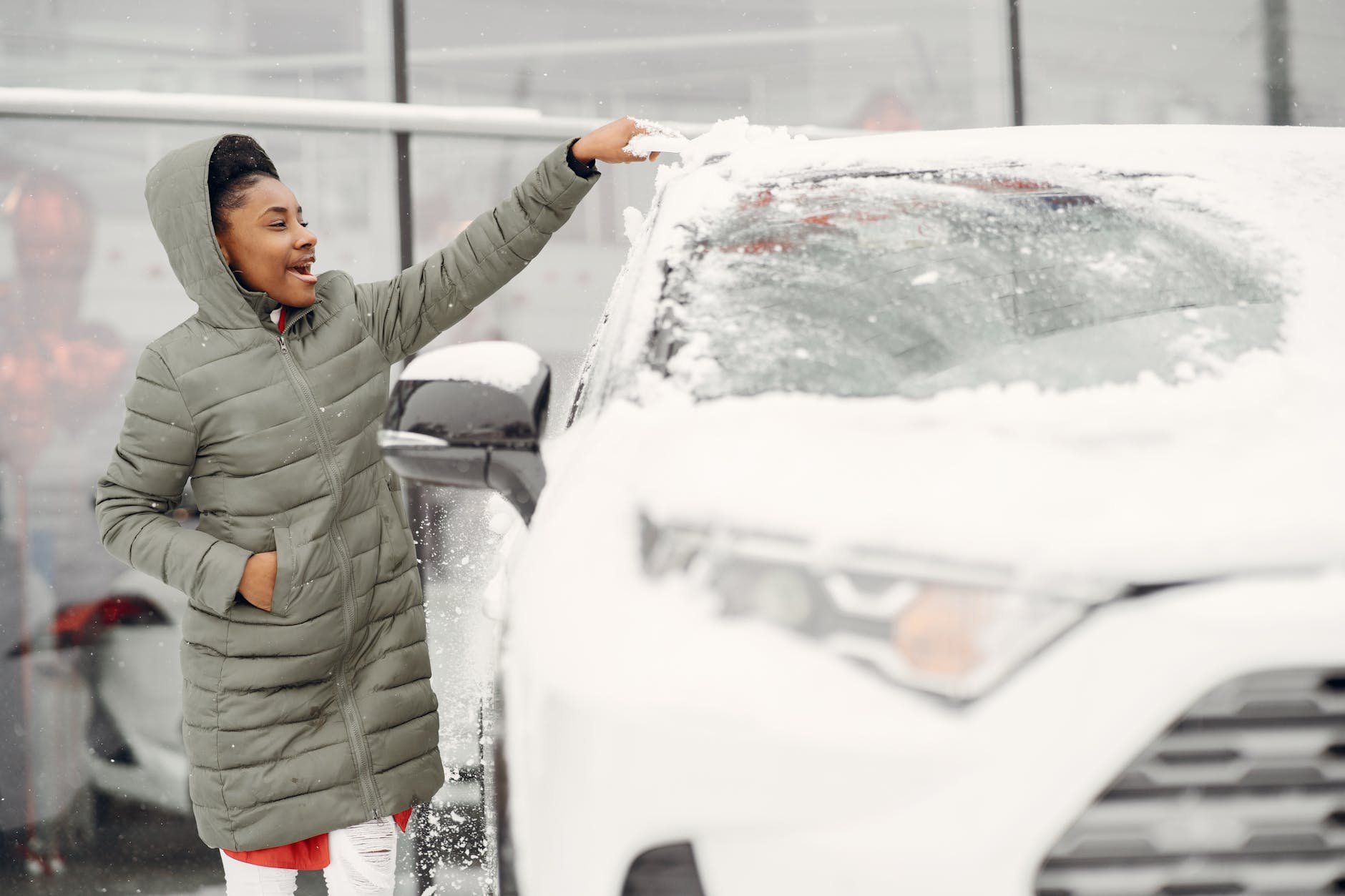 Smiling woman in winter coat brushing snow off her car's windshield outdoors.