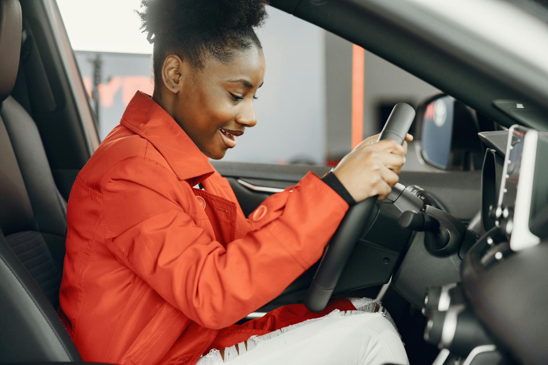 Young woman in an orange jacket enthusiastically trying out a car's steering wheel.